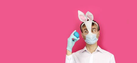 banner with man in mask Easter Bunny and medical mask look to blue egg, virus or coronavirus protection and prevention before holiday, preparation for party, copy space with pink backgroundの写真素材