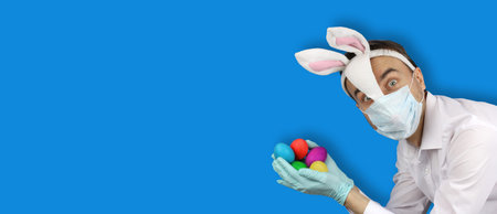 man in easter bunny and medical mask hold colored eggs, holidays in quarantine, virus or coronavirus...