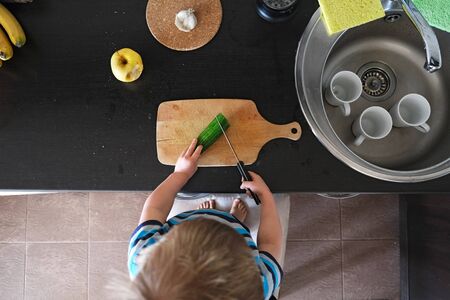 small vegetarian child cuts big green cucumber with knife, vegan kid helper cooks vegetable salad in kitchen, montessori training skill of coordination and independence, top view aboveの写真素材