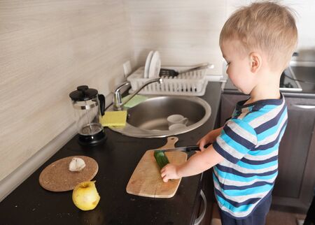 alone little vegetarian child cuts big green cucumber with knife, vegan kid helper cooks vegetable salad in kitchen, montessori training skill of coordination and independence, side viewの写真素材