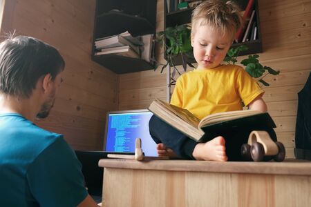 father freelancer working from home in quarantine, child sits near and read book, concept of self-isolated lifestyle home office workspace workplace, parenthood multitaskingの写真素材