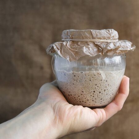 baker hand holds glass jar with fresh rye sourdough on whole grain flour on brown sackcloth background, yeast-free leaven starter for healthy organic rustic bread, square frameの写真素材