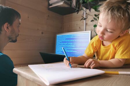 father freelancer developer working from home in quarantine, son child lying near and draws, concept of self-isolated lifestyle workspace workplace, parenthood multitaskingの写真素材