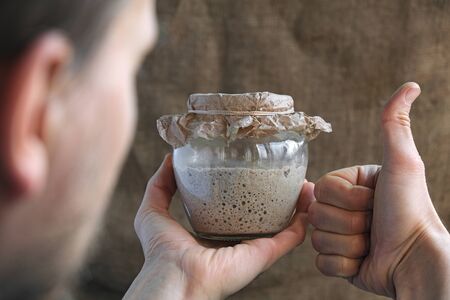 baker hold in hand glass jar with fresh rye sourdough on whole grain flour on brown sackcloth background and make thumb up gesture, like sign, starter for healthy organic bread, view from behindの写真素材