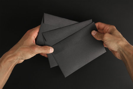 two caucasian male hands holding three blank black envelopes on black background, mockup with copy...
