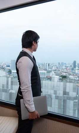 A young businessman monitors and keeps track of tasks in his office with a cityscape in the background of his desk.の写真素材