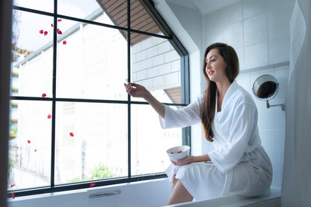 Woman sitting on bathtub wearing white bathrobe in modern bathroom at home with rose leaves to...