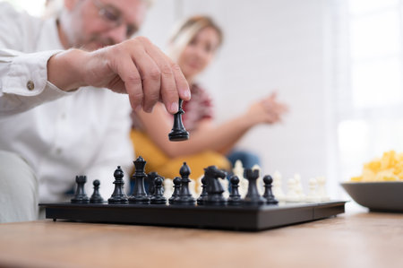 In the living room of the house, an elderly couple sits and relaxes. to begin playing chess together with a chess board with a daughter cheering beside himの写真素材