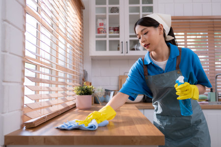 Cleaning service. Close-up of woman in apron and rubber gloves cleaning wooden table with sprayの写真素材