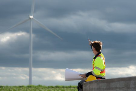 Young engineer working on a wind turbine field looking at the blueprint in the middle of the black clouds with sunlight shining, The concept of natural energy from wind.の写真素材