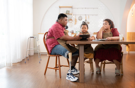 A plus-size family with a father wearing a prosthetic leg, is happily assisting a child with her homework and having fun together in the dining room of the house before cooking togetherの写真素材