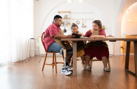 A plus-size family with a father wearing a prosthetic leg, is happily assisting a child with her homework and having fun together in the dining room of the house before cooking togetherの写真素材