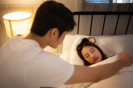 Young Asian man covers his wife with a blanket as she sleeps on the bed.の写真素材
