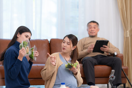 A pregnant asian woman sits on the floor with her younger sister. Live social media broadcast selling pregnant mothers' products and meals, There is a father in the background relaxing on a sofa.の写真素材