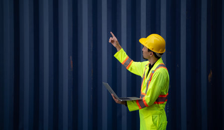 Workers in the import and export industry holding a laptop and standing on the front of container to...
