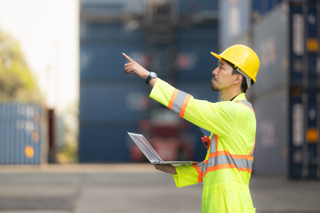 Workers in the import and export industry holding a laptop and standing on the front of container to...