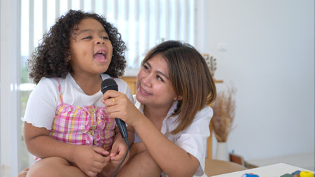 A tiny girl and mother sits on a toy table, clutching a microphone, and sings cheerfully in the living roomの写真素材