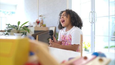 A tiny girl sits on a toy table, clutching a microphone, and sings cheerfully in the living roomの写真素材