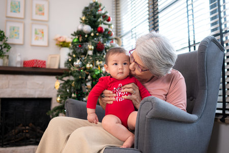 Grandma is sitting in an armchair, holding her infant grandson, playfully chatting with him in the...