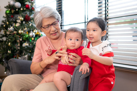 Grandma is sitting in an armchair, holding her infant grandson, playfully chatting with him in the living room, which is decorated for the upcoming Christmas celebrationの写真素材
