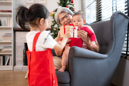 Grandma is sitting in an armchair, holding her infant grandson, playfully chatting with him in the living room, which is decorated for the upcoming Christmas celebrationの写真素材