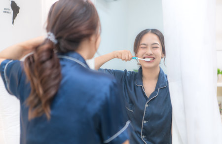 Asian young woman in a blue pajama is brushing her teeth in front of the mirror before taking a...