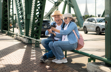An elderly couple relaxes and looks at photos in camera after walking through the city streets with the road and bridge structures in the backgroundの写真素材