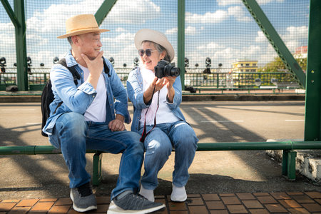 An elderly couple relaxes and looks at photos in camera after walking through the city streets with the road and bridge structures in the backgroundの写真素材