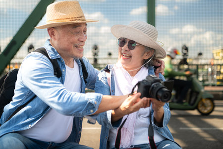 An elderly couple relaxes and looks at photos in camera after walking through the city streets with the road and bridge structures in the backgroundの写真素材