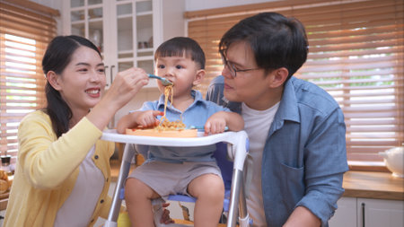 Happy Asian parents enjoying mealtime with their toddler in the kitchen, sharing laughter and playful moments during feeding timeの写真素材
