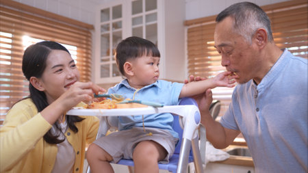 Happy Asian parents enjoying mealtime with their toddler in the kitchen, sharing laughter and playful moments during feeding timeの写真素材