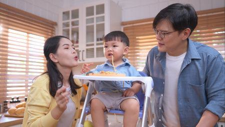 Happy Asian parents enjoying mealtime with their toddler in the kitchen, sharing laughter and playful moments during feeding timeの写真素材