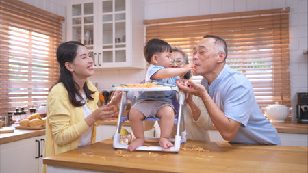 Happy Asian parents enjoying mealtime with their toddler in the kitchen, sharing laughter and playful moments during feeding timeの写真素材