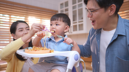 Happy Asian parents enjoying mealtime with their toddler in the kitchen, sharing laughter and playful moments during feeding timeの写真素材