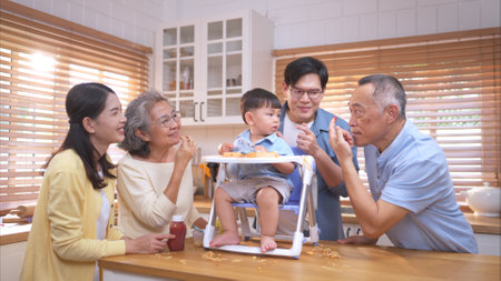 Happy Asian parents enjoying mealtime with their toddler in the kitchen, sharing laughter and playful moments during feeding timeの写真素材