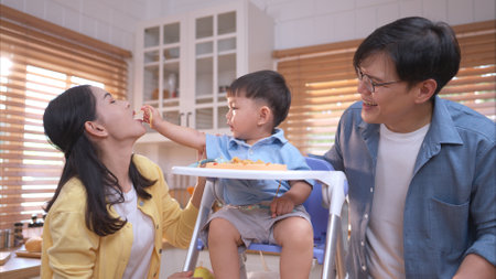 Happy Asian parents enjoying mealtime with their toddler in the kitchen, sharing laughter and playful moments during feeding timeの写真素材