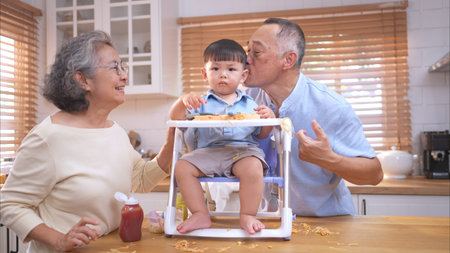 Happy Asian parents enjoying mealtime with their toddler in the kitchen, sharing laughter and...