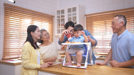 Happy Asian parents enjoying mealtime with their toddler in the kitchen, sharing laughter and...