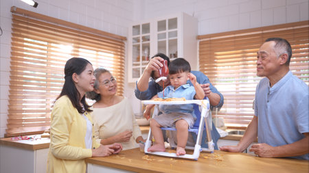 Happy Asian parents enjoying mealtime with their toddler in the kitchen, sharing laughter and playful moments during feeding timeの写真素材