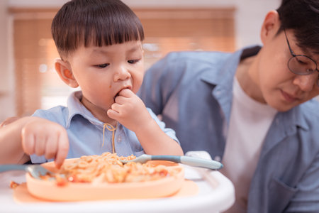 Happy Asian parents enjoying mealtime with their toddler in the kitchen, sharing laughter and playful moments during feeding timeの写真素材