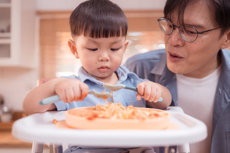 Happy Asian parents enjoying mealtime with their toddler in the kitchen, sharing laughter and playful moments during feeding timeの写真素材