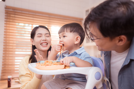 Happy Asian parents enjoying mealtime with their toddler in the kitchen, sharing laughter and playful moments during feeding timeの写真素材
