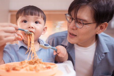 Happy Asian parents enjoying mealtime with their toddler in the kitchen, sharing laughter and playful moments during feeding timeの写真素材
