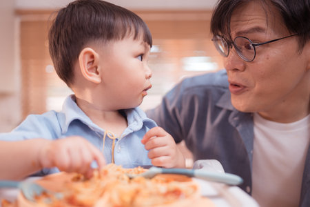 Happy Asian parents enjoying mealtime with their toddler in the kitchen, sharing laughter and playful moments during feeding timeの写真素材