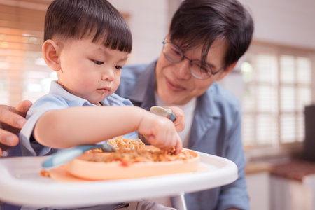 Happy Asian parents enjoying mealtime with their toddler in the kitchen, sharing laughter and playful moments during feeding timeの写真素材