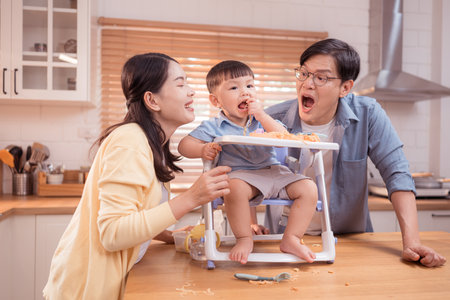 Happy Asian parents enjoying mealtime with their toddler in the kitchen, sharing laughter and playful moments during feeding timeの写真素材