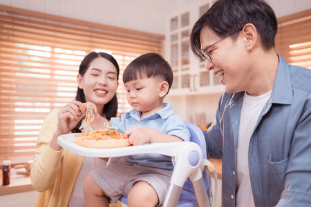 Happy Asian parents enjoying mealtime with their toddler in the kitchen, sharing laughter and playful moments during feeding timeの写真素材