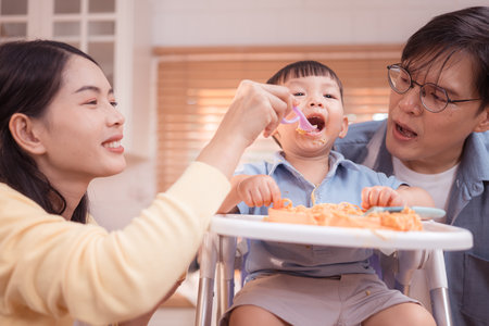 Happy Asian parents enjoying mealtime with their toddler in the kitchen, sharing laughter and playful moments during feeding timeの写真素材