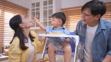Happy Asian parents enjoying mealtime with their toddler in the kitchen, sharing laughter and playful moments during feeding timeの写真素材