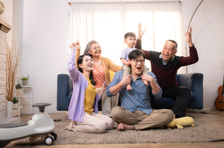 Happy multi-generational Asian family enjoying quality time together at home, cheering and smiling with a toddler riding a toy car in a cozy living roomの写真素材